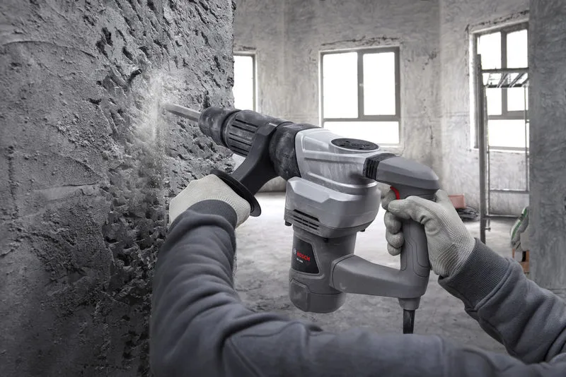 A person wearing safety equipment uses a demolition hammer to break a concrete wall.