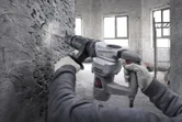 A person wearing safety equipment uses a demolition hammer to break a concrete wall.