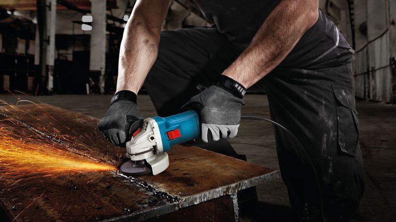 A person wearing safety equipment grinds a metal beam with a power tool, creating sparks.