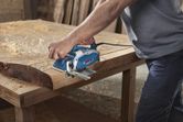 A person uses an electric planer to smooth a wooden plank on a workbench.