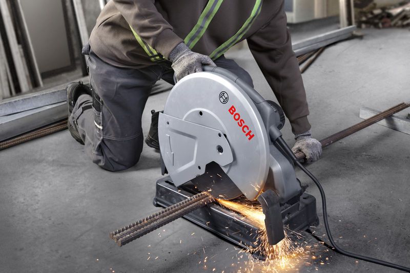 A person wearing safety equipment cuts metal rebar with a chop saw, creating sparks.