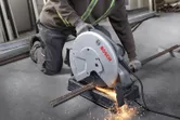 A person wearing safety equipment cuts metal rebar with a chop saw, creating sparks.