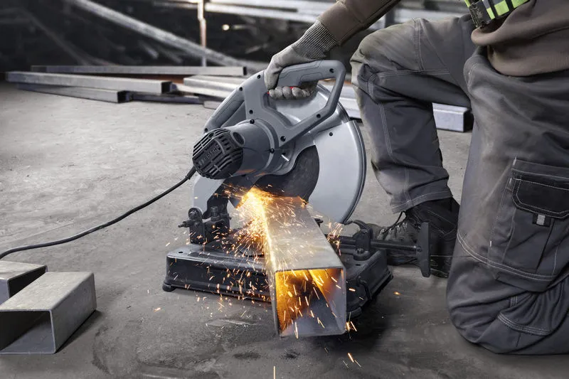 A person wearing safety equipment cuts metal tubing with a chop saw, sparks flying.