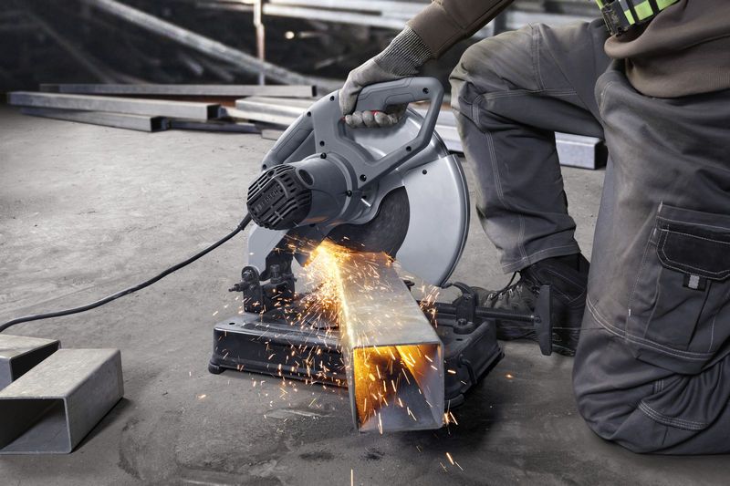 A person wearing safety equipment cuts metal tubing with a chop saw, sparks flying.