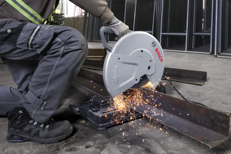 A person wearing safety equipment operates a chop saw cutting a steel beam.