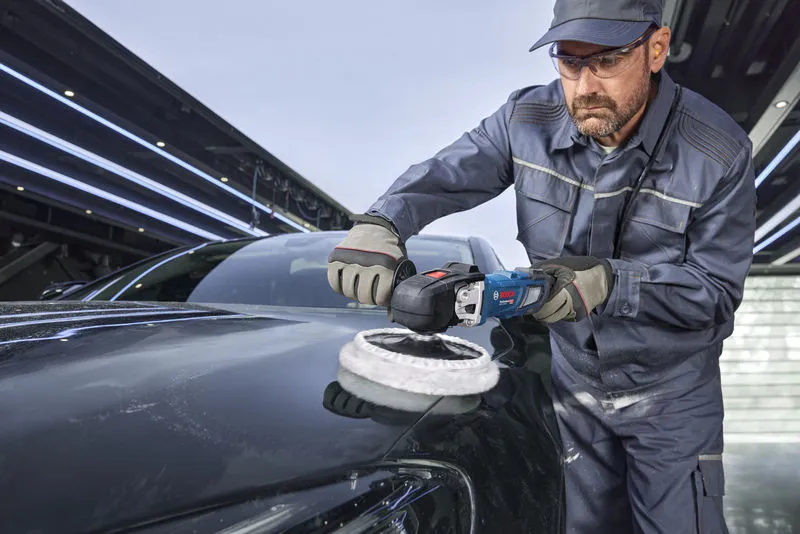 A person wearing safety equipment polishes a black car hood with a power polisher.