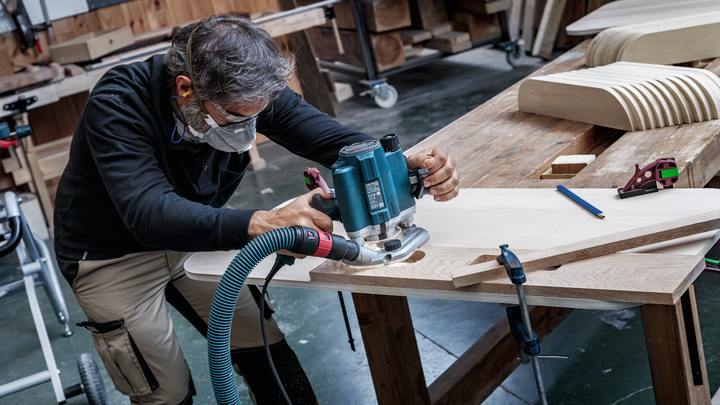 Person wearing safety equipment shapes wood using a power router in a workshop.