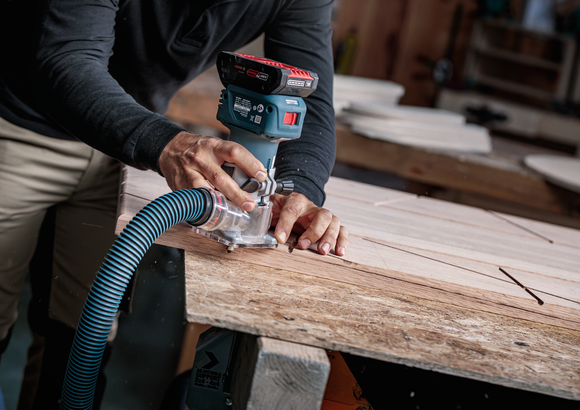 Person guides cordless router along wooden board in a workshop.