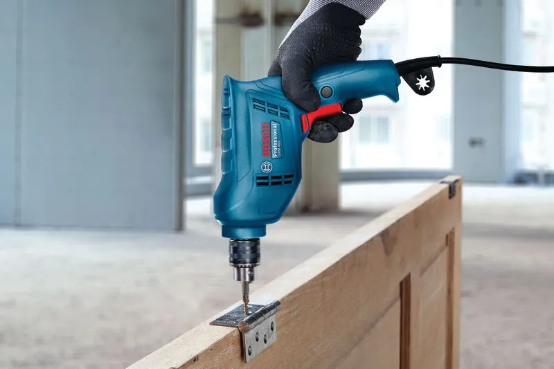 A person wearing safety equipment uses a power drill to install a hinge on a wooden door.