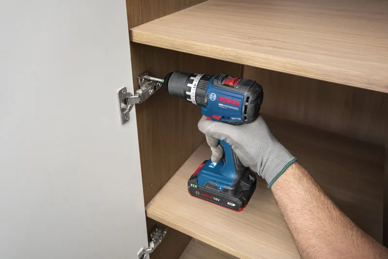 A person wearing safety equipment uses a cordless drill to install a cabinet hinge.