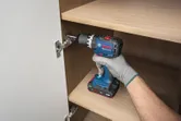 A person wearing safety equipment uses a cordless drill to install a cabinet hinge.