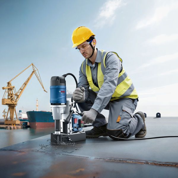 Person wearing safety equipment operates a magnetic core drill on a metal surface at a shipyard.