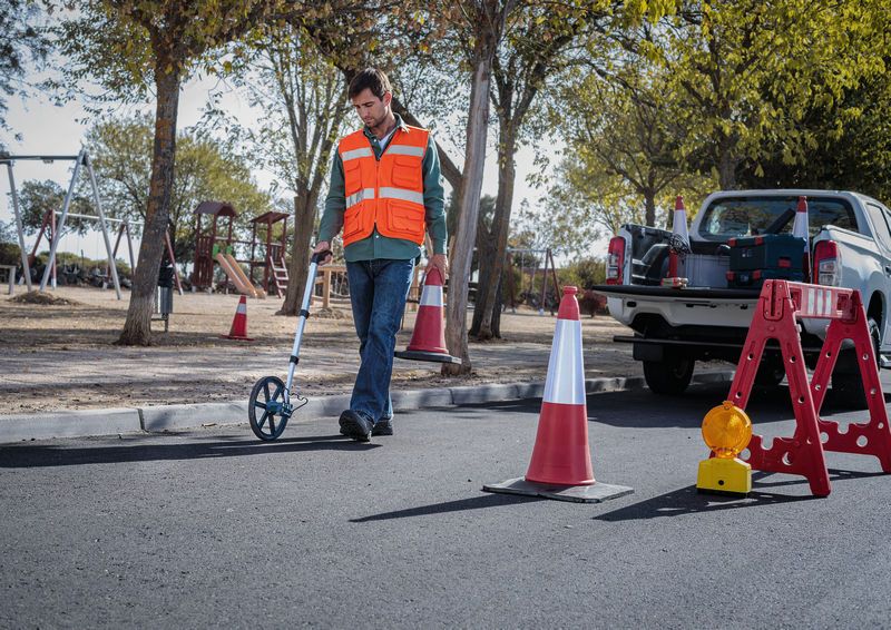 A person wearing safety equipment measures road distance using a measuring wheel.