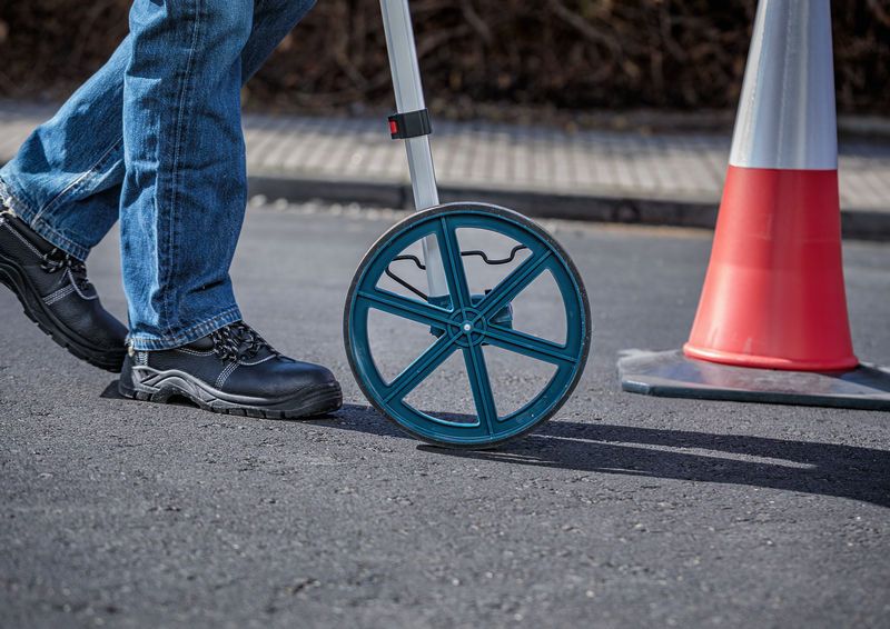A person wearing safety equipment uses a measuring wheel on a paved surface.