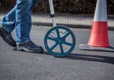 A person wearing safety equipment uses a measuring wheel on a paved surface.