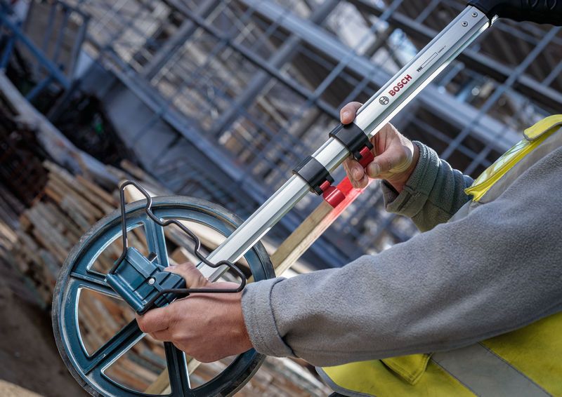 A person wearing safety equipment measures distance with a large measuring wheel.