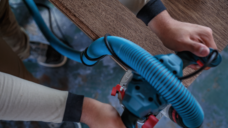 Person guides a power tool with a blue vacuum hose along the edge of a wooden board.