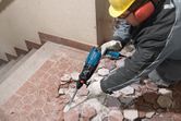 A person wearing safety equipment uses a demolition tool to break floor tiles.