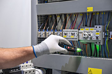 A hand wearing a white glove holds a green Bosch Professional voltage tester near electrical components. Wires in various colors run along a gray panel with labeled switches and circuit breakers, indicating an electrical setup. The background is simple and neutral, emphasizing the tools and components.