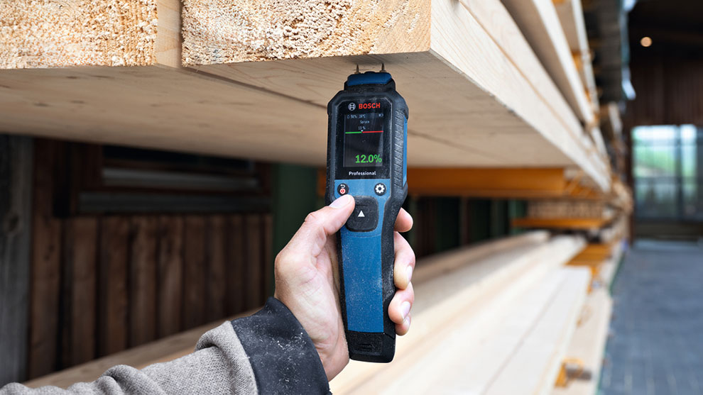 A person holds a Bosch Professional moisture meter, displaying 12.0%, beside stacked wooden planks.