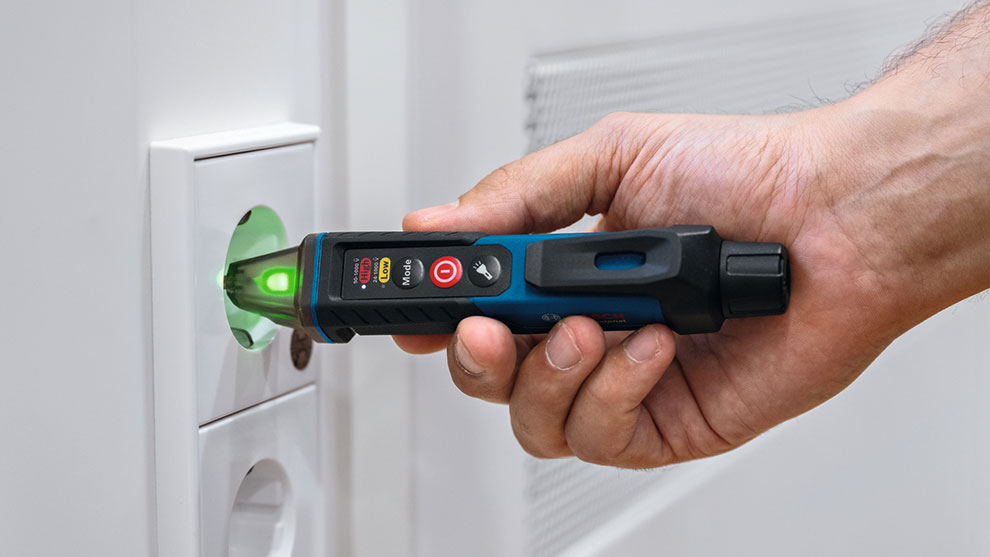 A hand holds a Bosch Professional voltage tester, with a green light indicating power detection, near a wall socket.