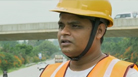 A man in a yellow hard hat and safety vest stands on a road, with a bridge in the background.