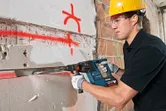 A person wearing safety equipment chisels a groove in a wall with a rotary hammer.