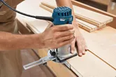 A person uses a palm router to shape the edge of a wooden board.
