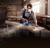 A person wearing safety equipment smooths a wooden plank with a planer.
