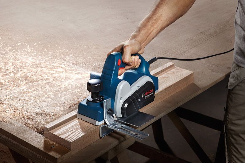 A person uses an electric planer to smooth a wooden board on a workbench.