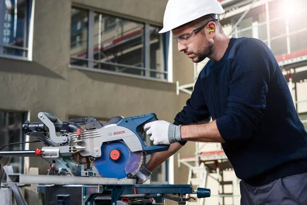 Worker wearing safety equipment cuts wood with a circular saw at a construction site.