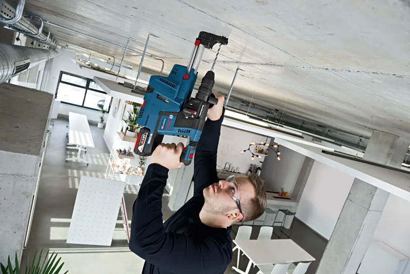 A person wearing safety equipment drills into a concrete ceiling in a modern workspace.