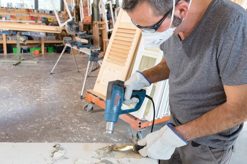 A person wearing safety equipment uses a heat gun to remove paint from a wooden surface.