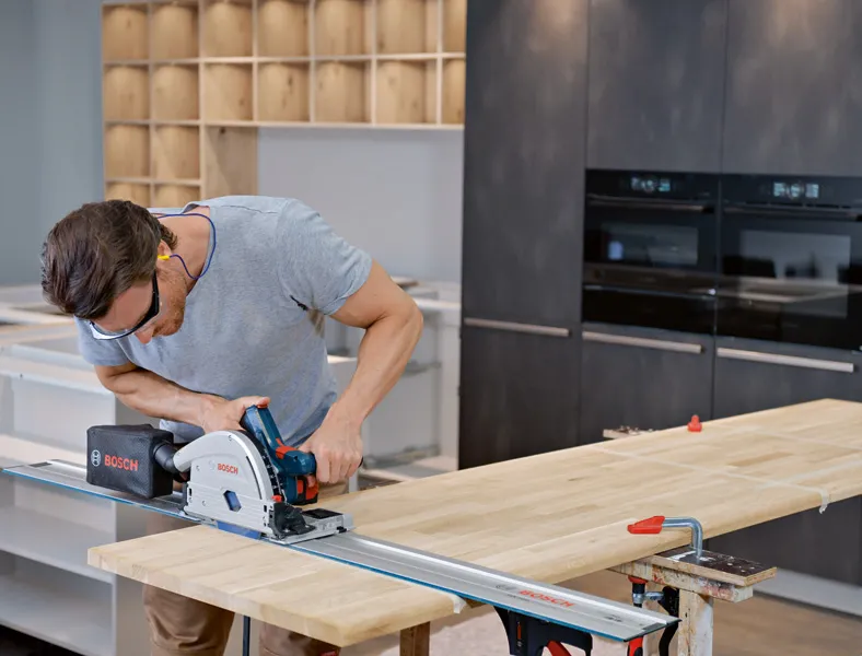 Person wearing safety equipment cuts a wooden board with a track saw in a modern kitchen.
