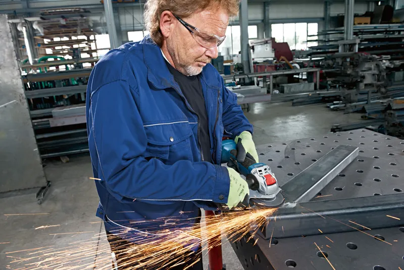A person wearing safety equipment grinds metal with a cordless angle grinder.