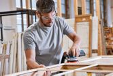 A person wearing safety equipment sands wood with a power sander in a workshop.
