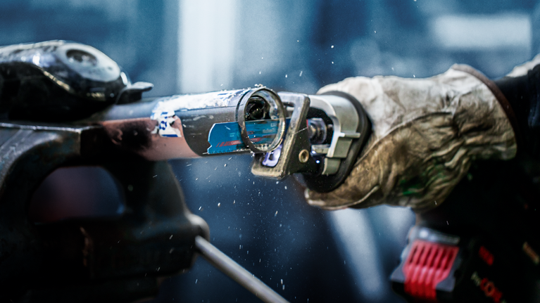 Person wearing safety equipment uses a reciprocating saw to cut through a metal pipe.