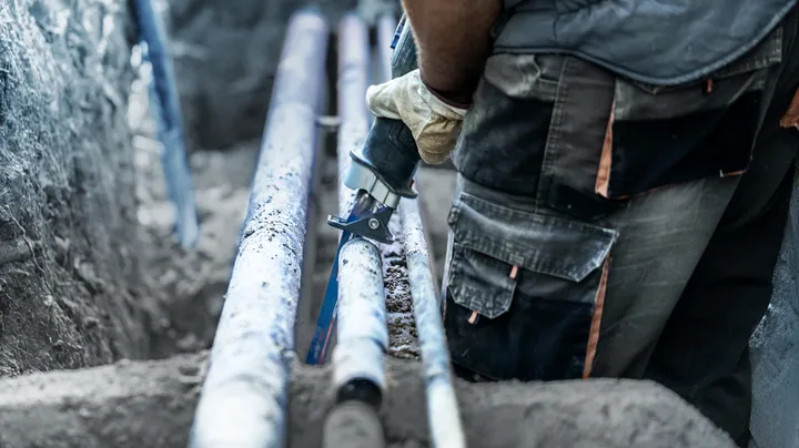 Person wearing safety equipment cuts a pipe with a power saw in a trench.