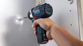 A person uses a cordless impact driver to drive a screw into a white surface.
