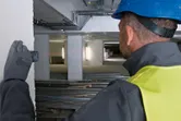 A person wearing safety equipment uses a laser measure to gauge distance in a construction area.