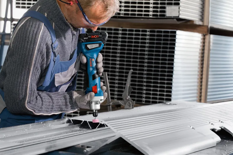 A person wearing safety equipment grinds a metal sheet with a cordless straight grinder.