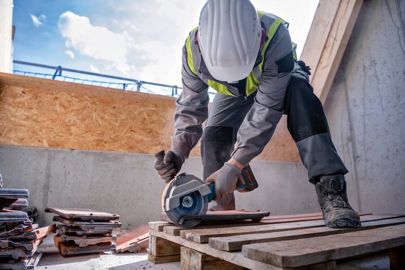 A person wearing safety equipment cuts roof tiles with a cordless angle grinder.