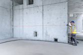A person wearing safety equipment measures a concrete wall with a laser measure.