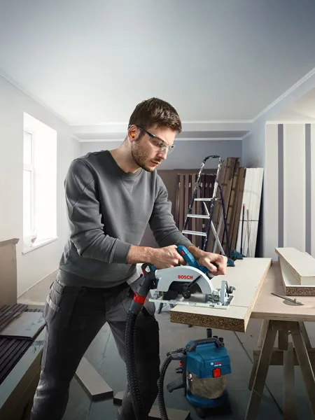 Person wearing safety equipment cuts wood with a cordless circular saw in a workshop.