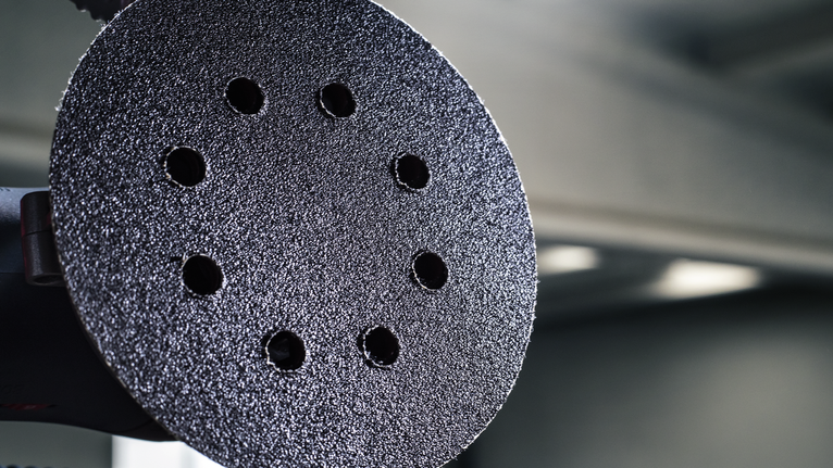 Close-up of a circular sanding pad with multiple holes against a blurred workshop background.