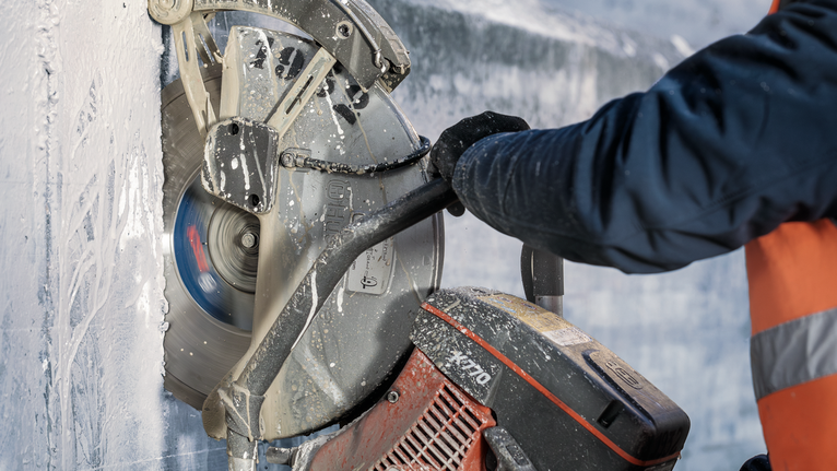 A person wearing safety equipment uses a large saw to cut through thick concrete.