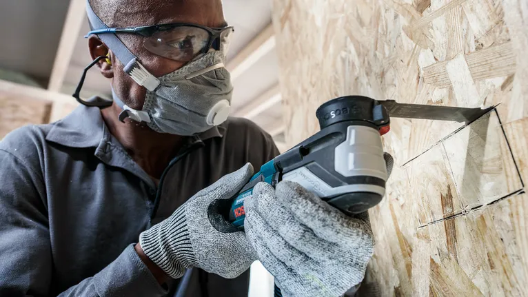 Person wearing safety equipment uses a power tool to cut a square opening in wood.