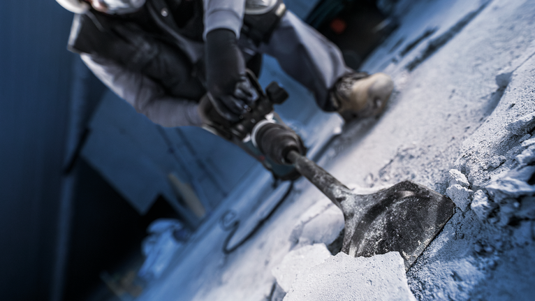 A person wearing safety equipment breaks concrete with a demolition tool.