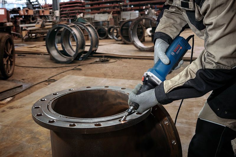 A person wearing safety equipment grinds the edge of a large metal cylinder with a straight grinder.