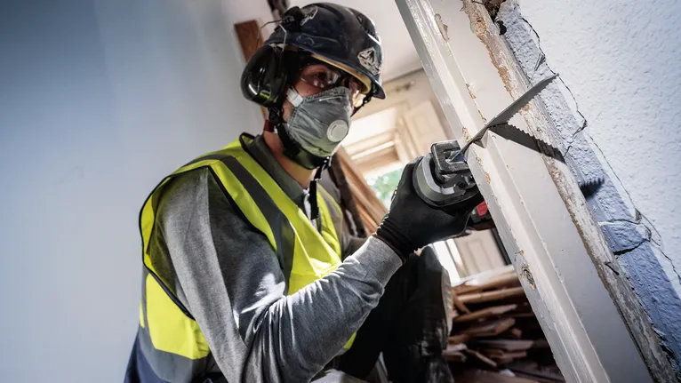 Person wearing safety equipment cuts a doorframe with a reciprocating saw.